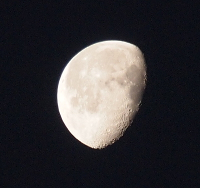 [A three-quarter moon missing the right section from about 1 o'clock to 7 o'clock has clearly visible craters in the lower right portion as well as light and dark grey sections on its surface in this close view image. The background sky is a deep navy blue.]